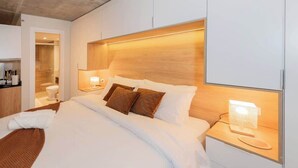 Quarto de hotel moderno com uma cama grande, abajures e uma cabeceira de madeira.