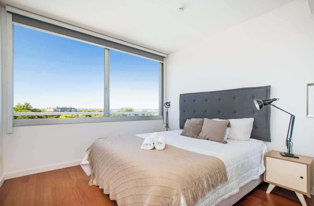 Ein Schlafzimmer mit großem Fenster, einem Bett mit grauem Kopfteil, einem Nachttisch und einer Lampe.