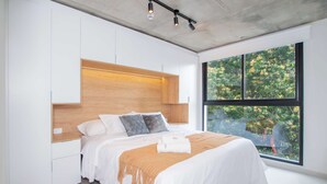 Ein modernes Schlafzimmer mit einem großen Bett, einem festen Holz-Kopfstück, weißen Wänden und einem großen Fenster mit Blick auf Grünflächen.