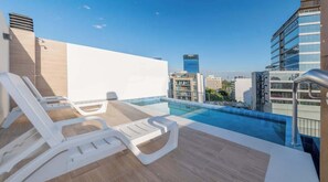 Ein Pool auf dem Dach mit Liegestühlen, freier Blick auf die Stadtlandschaft und moderne Gebäude im Hintergrund.