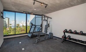Ein Fitnessraum mit Laufband, Gewichtbank und Kurzhanteln, großen Fenstern mit Blick auf die Stadt und einer Beton Decke.