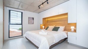 A modern bedroom with a large bed, bedside lamps, a wooden headboard, and a view through a sliding door. - Modern 1BR Near Shopping del Sol | Pool & Gym (Asunción)