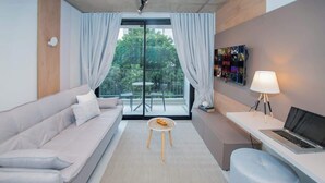 Una camera moderna con un divano, un tavolino rotondo, una scrivania con un portatile, una TV a parete e un balcone con vista sul verde.