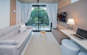 Quarto moderno com sofá, mesa redonda pequena, escrivaninha com laptop, TV fixada na parede e varanda com vista para vegetação.