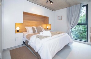 Quarto com cama, luminárias laterais, janela com cortinas e vista para o verde lá fora.