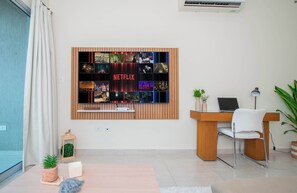Ein modernes Wohnzimmer mit an der Wand montiertem Fernseher, der eine Netflix-Oberfläche zeigt, einem Holztisch, einem weißen Stuhl, einer kleinen Pflanze und einer Lampe.