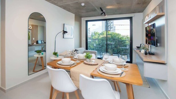 Un'area pranzo moderna con tavolo in legno apparecchiato per quattro persone, sedie bianche e una grande finestra con vista su edifici e vegetazione.