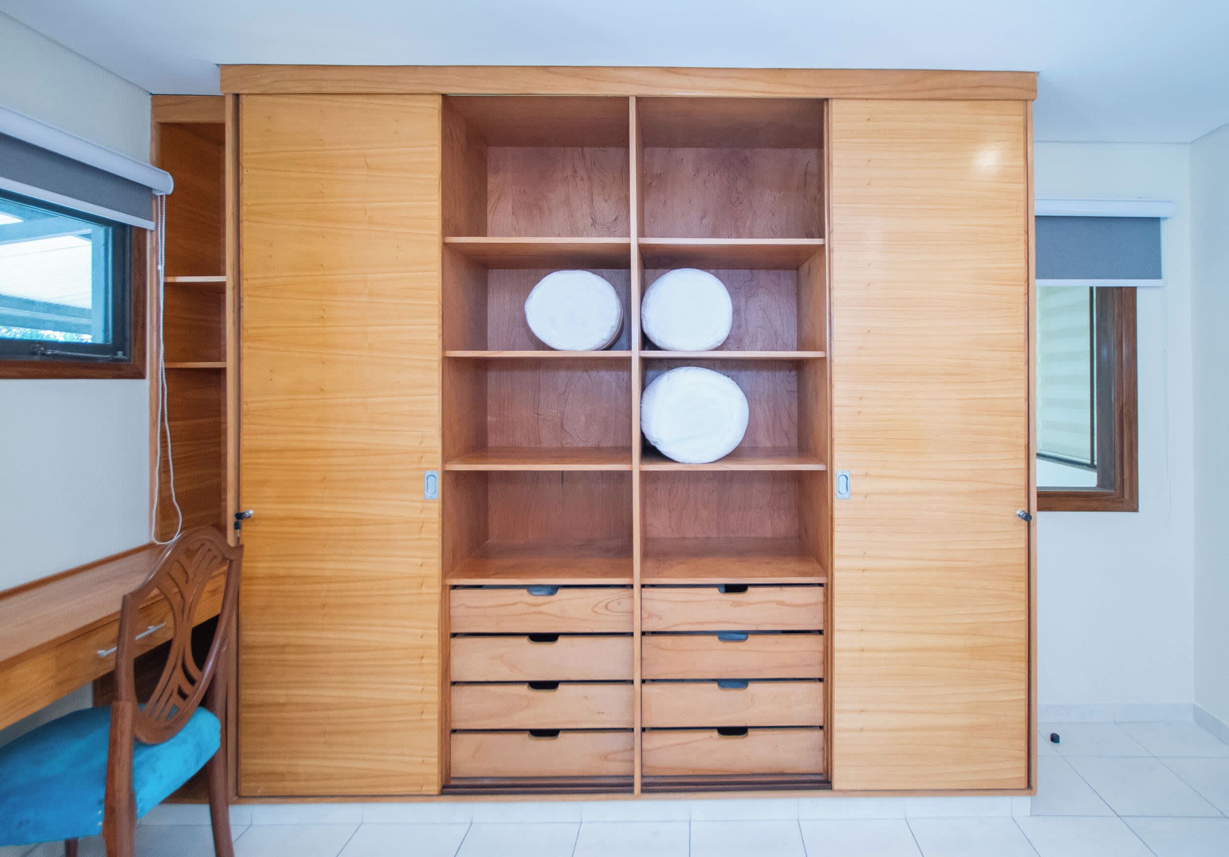 A wooden wardrobe with multiple shelves and drawers, a desk with a chair, and a window.