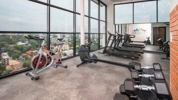 A gym with treadmills, stationary bikes, and dumbbells, large windows with a city view, and a sign reading 'Elite Fitness'.