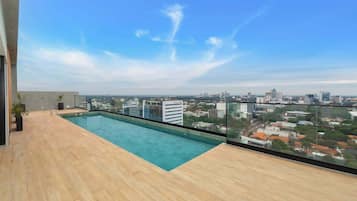 A rooftop pool with a city view, wooden deck, and a glass railing.