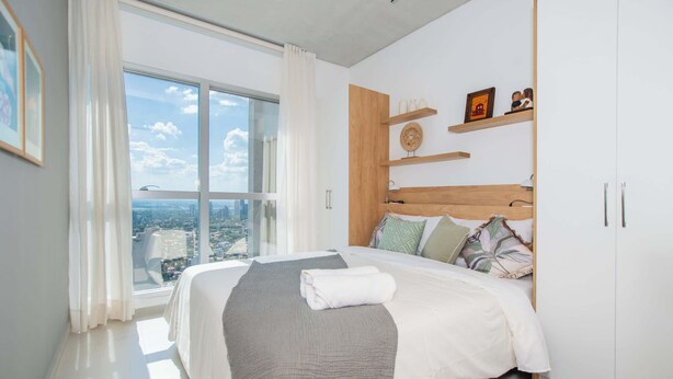 A modern bedroom with a large window, a bed with a white and gray blanket, wooden headboard, and a city view. - Modern One BR in Petra Tower, Skyline Views & Pool (Asunción)