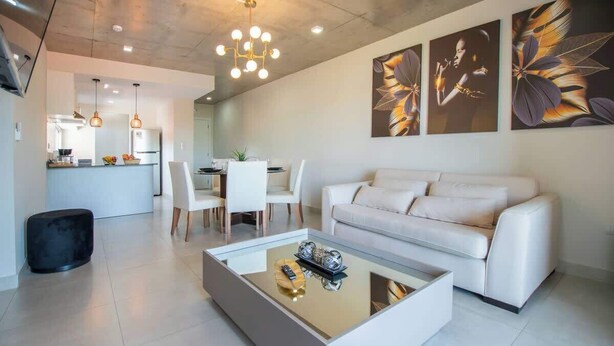 A modern living room with a glass coffee table, a white sofa, and a dining area with a white table and chairs.
