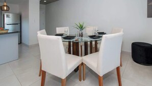 A dining area with a round glass table, white upholstered chairs, and a modern kitchen in the background.