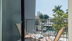 Een balkon met een glazen deur, metalen stoelen, een kleine tafel en uitzicht op een straat met gebouwen en bomen.