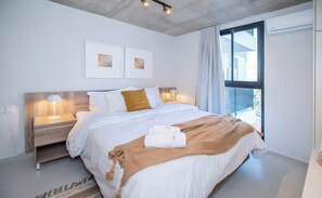 Quarto de hotel moderno com uma cama grande, duas mesas de cabeceira com abajures, vista para o exterior e obras de arte na parede.
