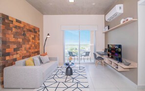 Una sala de estar moderna con pared de ladrillo, un sofá, una mesa de centro, un televisor y un balcón con sillas.