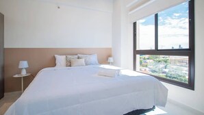 A bedroom with a large bed, two bedside tables with lamps, a view through a large window, and a cityscape outside.