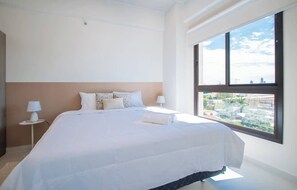 Un dormitorio con una cama grande, dos mesitas de noche con lámparas, vistas a través de un ventanal y un paisaje urbano al exterior.