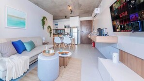 Un soggiorno moderno con divano, tavolino e poltrone, una cucina sullo sfondo e una TV a muro che mostra Netflix.