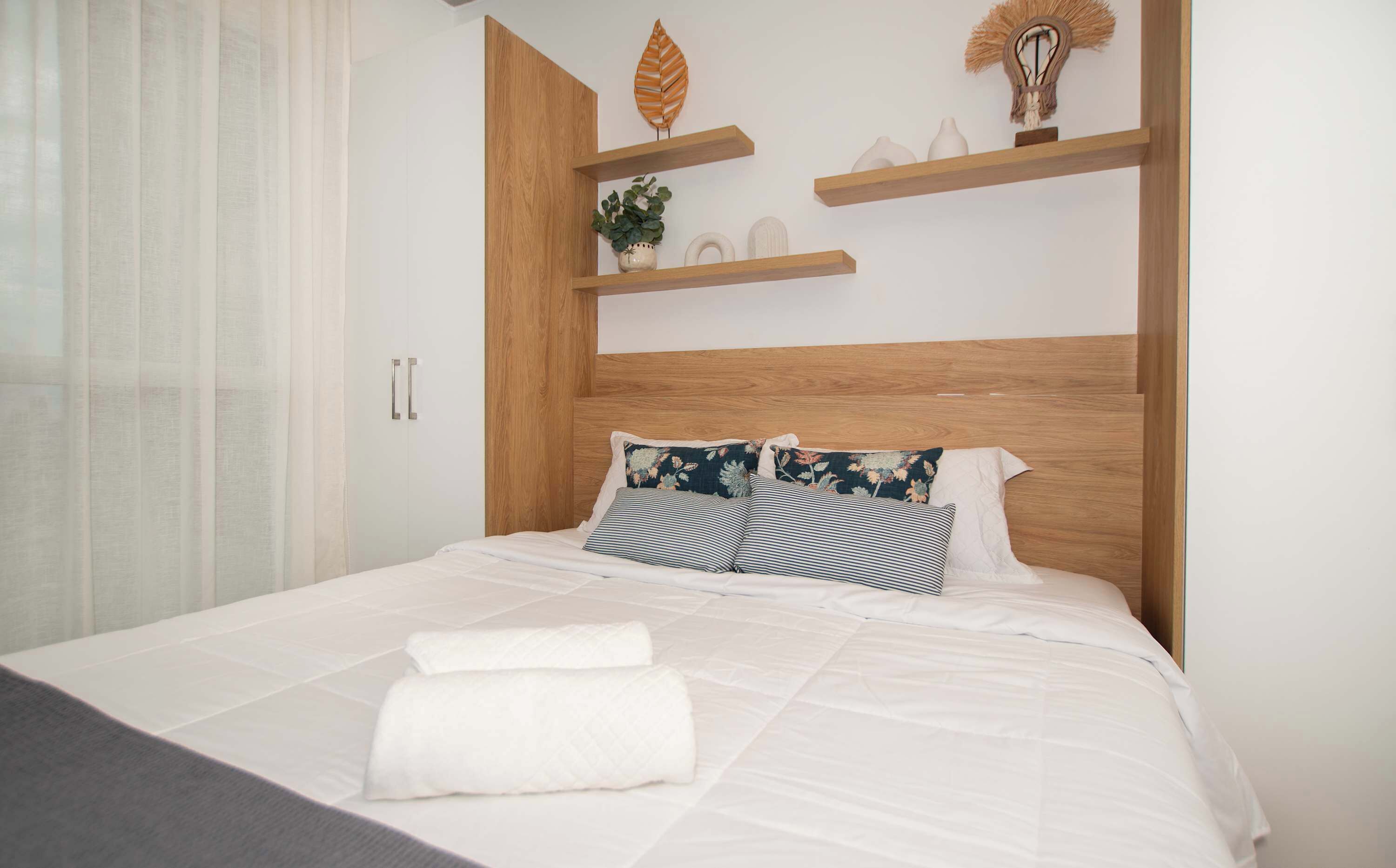 Un lit bien fait, avec du linge de lit blanc et des oreillers à rayures, une tête de lit en bois et une étagère de chevet agrémentée d’objets décoratifs.