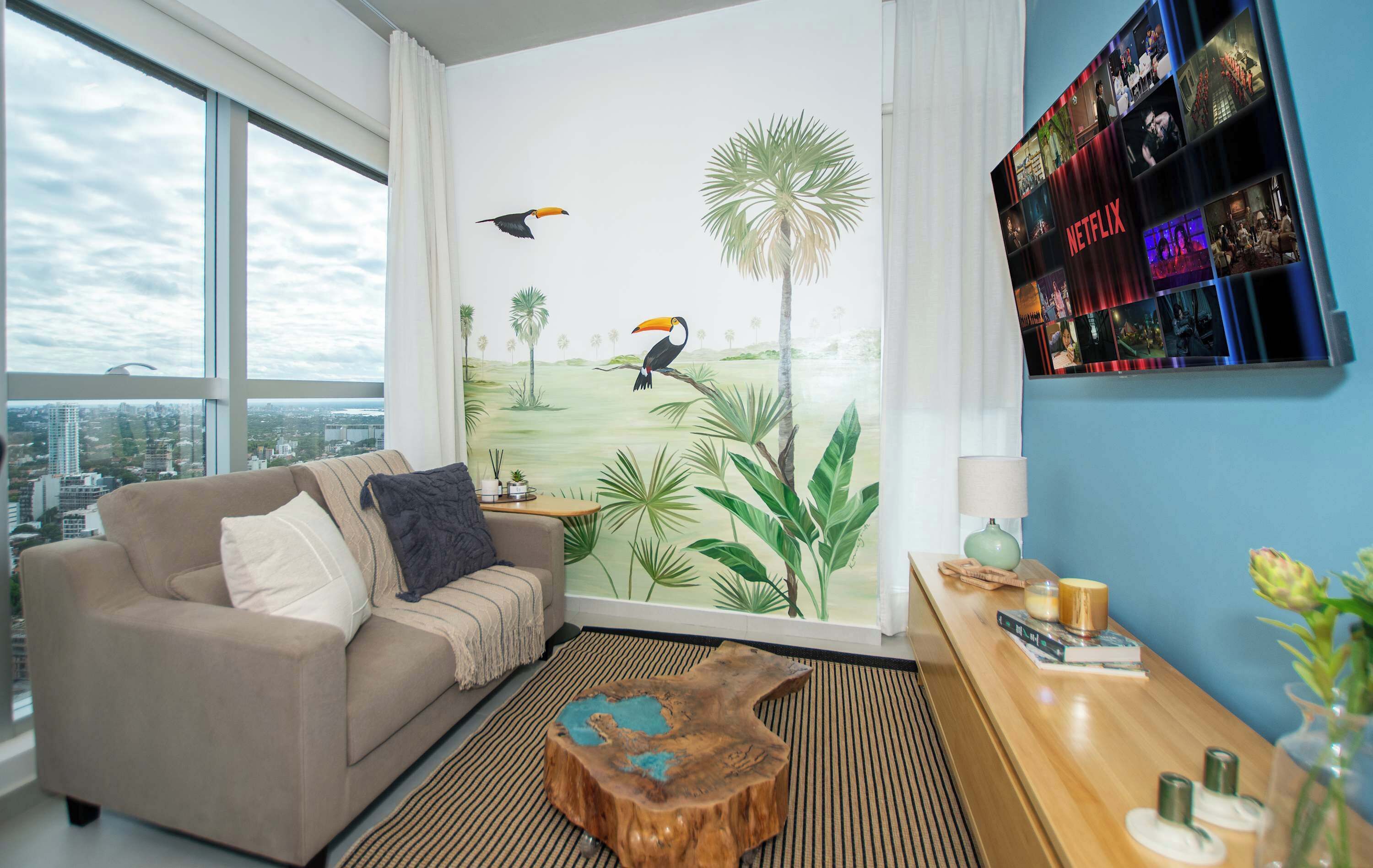 Un salon doté d’une grande fenêtre, d’un canapé, d’une table basse, d’une fresque murale et d’un écran fixé au mur.