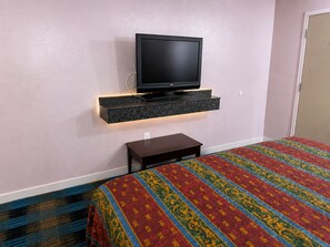 A hotel room with a flat-screen TV mounted on a black shelf, a small bedside table, and a patterned bedspread.