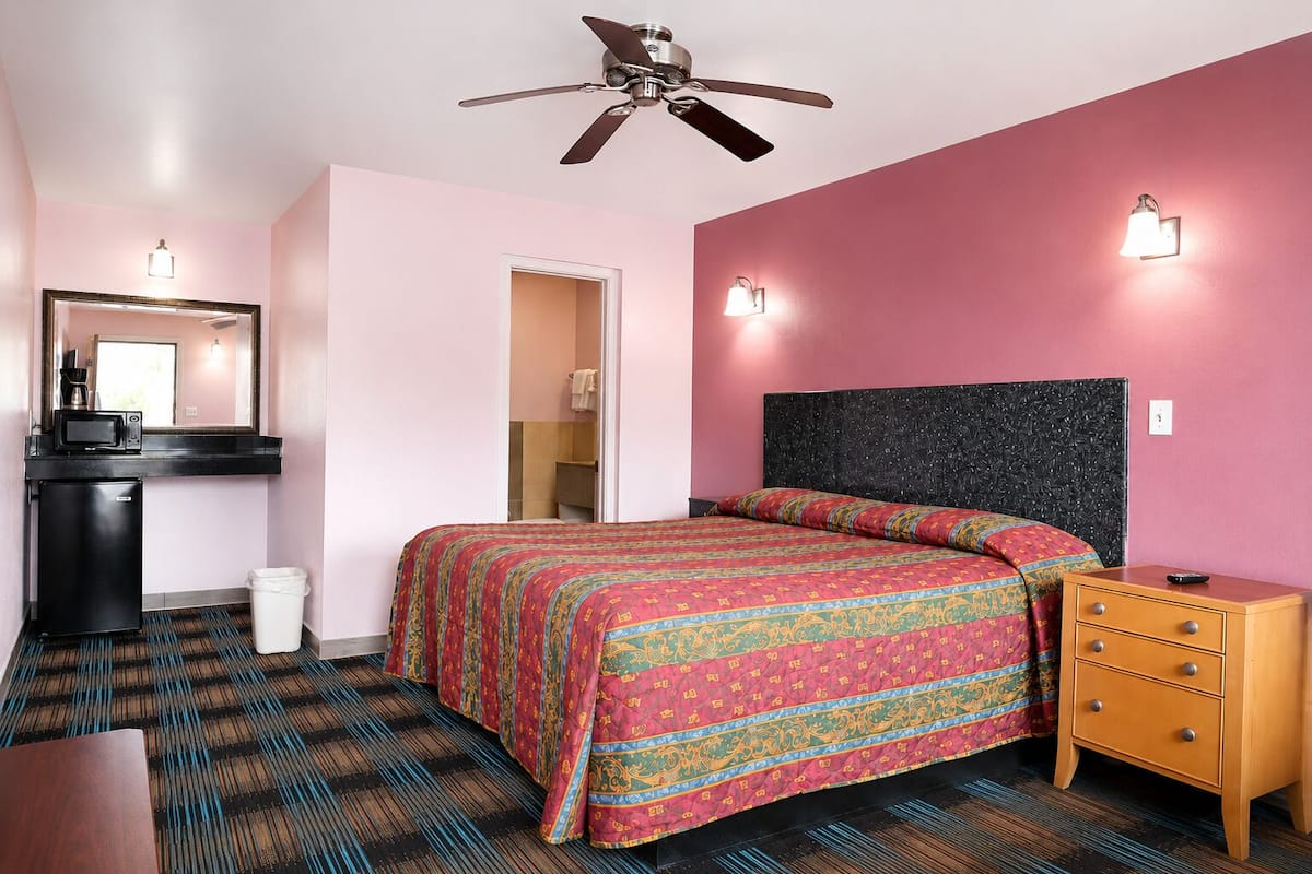 A bedroom with a bed, a dresser, a desk with a microwave, a trash bin, a ceiling fan, and a bathroom door.