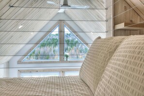 A bedroom with a sloped ceiling, a ceiling fan, a large window with a view of trees, and a bed with patterned bedding. - Peaceful Lakeside Cabin Retreat with Beach, Fire Pit & Kayaks (Sainte-Marguerite-du-Lac-Masson)