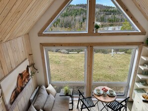 A room with a lofted ceiling, a sofa, a dining table with chairs, and a view of a grassy field and forest. - Peaceful Lakeside Cabin Retreat with Beach, Fire Pit & Kayaks (Sainte-Marguerite-du-Lac-Masson)