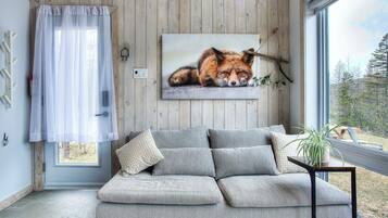 Une pièce chaleureuse avec un canapé gris, une fenêtre donnant sur des arbres et un tableau encadré représentant un renard accroché au mur.