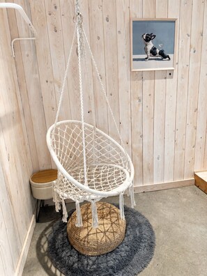 A white macrame hanging chair with a woven base, a small wooden stool, and a framed picture of a dog on a wooden wall. - Peaceful Lakeside Cabin Retreat with Beach, Fire Pit & Kayaks (Sainte-Marguerite-du-Lac-Masson)