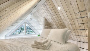 Un dormitorio con techo inclinado, una cama con manta texturizada, almohadas y una ventana con vistas a árboles.