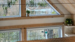 Una habitación con techo inclinado, vigas de madera, ventilador de techo y ventanas amplias que ofrecen vistas a un bosque y un cuerpo de agua.