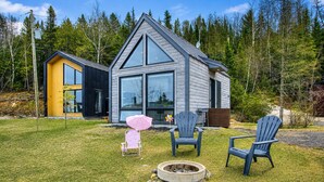Una cabaña moderna con un ventanal grande, una fogata y dos sillas Adirondack.