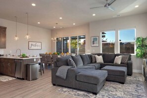 Ein modernes Wohnzimmer mit einem grauen Ecksofa, einer Küchenzeile mit Holzschränken, Pendelleuchten und großen Fenstern.