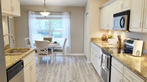 A kitchen with white cabinets, a dining table with chairs, a window with curtains, and a ceiling fan.