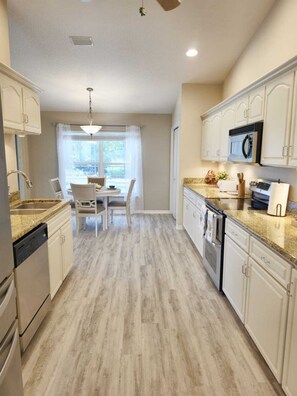 A kitchen with white cabinets, a dining table with chairs, a window with curtains, and a ceiling fan.
