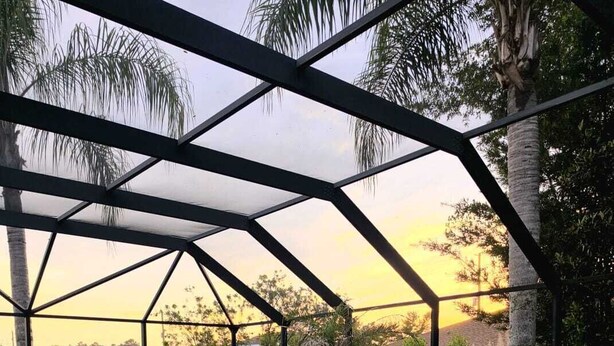 A swimming pool with a geometric metal roof structure, surrounded by palm trees and a grassy area.