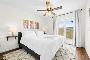 Ein Schlafzimmer mit einem Bett, Nachttischlampen, einem Deckenventilator, Bildern an der Wand und Blick durch ein Fenster.