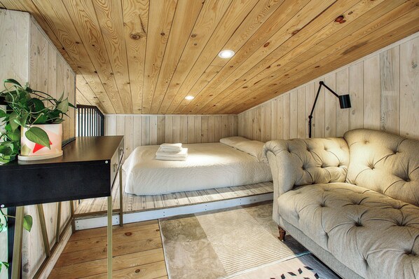 A bedroom with a wooden ceiling, a bed, a sofa, a desk with a plant, and a lamp. - Cozy Lakeside Cabin Retreat with Beach, Kayaks, Fire Pit & Trails (Sainte-Marguerite-du-Lac-Masson)