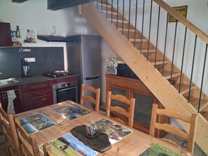 Eine kompakte Küche mit Gasherd, Backofen und Kühlschrank. Ein Holztisch mit Stühlen steht unter einer Treppe.