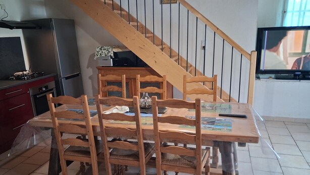 Spiseområde med et træbord og stole, en trappe, et fjernsyn samt et køkken med køleskab og komfur.