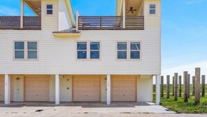 A two-story building with a flat roof, a balcony on the upper floor, and three garage doors. - Lux 2BR-2BA: Boardwalk + 3rd Floor Deck (Port Aransas)