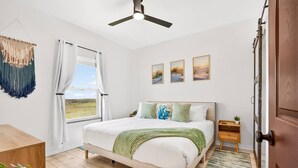 A bedroom with a bed, a ceiling fan, a window with curtains, a wooden door, and a small bedside table with a plant. - Lux 2BR-2BA: Boardwalk + 3rd Floor Deck (Port Aransas)