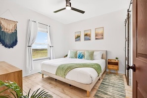 A bedroom with a bed, a ceiling fan, a window with curtains, a wooden door, and a small bedside table with a plant. - Lux 2BR-2BA: Boardwalk + 3rd Floor Deck (Port Aransas)
