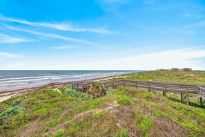 A coastal landscape with a wooden boardwalk leading to the beach, grassy terrain, and a clear view of the ocean. - Lux 2BR-2BA: Boardwalk + 3rd Floor Deck (Port Aransas)