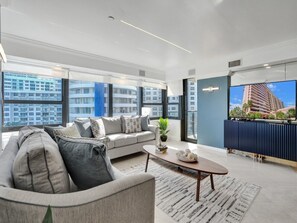 A modern living room with a sofa, coffee table, and a large window offering a city view. - Spacious Ocean View Condo w/ Beach Service, Pool, Gym 803 (Miami Beach)