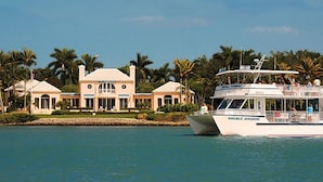 Uma casa grande de dois andares com fachada simétrica, um barco chamado 'DOUBLE SUNSHINE' atracado nas proximidades e palmeiras ao fundo.