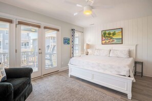 A bedroom with a white bed, a ceiling fan, a painting, and a view of the outside. - Wright Place Wright Time (Carolina Beach)