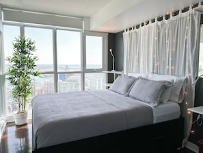 Un dormitorio con una cama grande, una planta en maceta y vistas de un paisaje urbano a través de amplios ventanales.